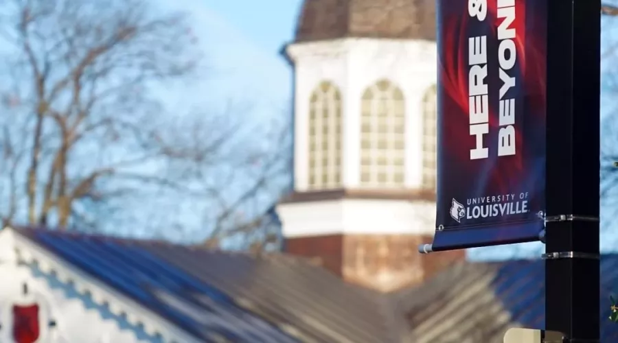 UofL's law school copula is out of focus in the background framed byt a lamppost banner reading "Here & Beyond" with UofL branding.